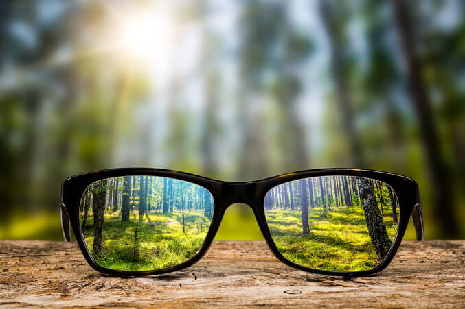 Brille auf einem Holztisch, durch die ein sonniger Wald scharf und klar zu sehen ist